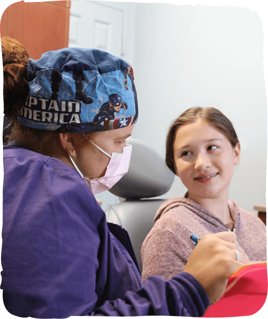 Pediatric dentist in blue mask and cap examines young girl's teeth in dental office.