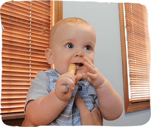Baby in blue shirt chewing on fingers in home setting with wooden blinds.
