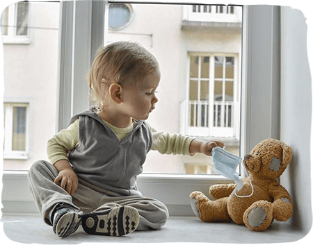 Young toddler sitting on windowsill reaching toward teddy bear, bright indoor setting.