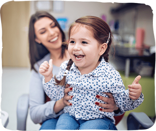 Young girl smiling at pediatric dental office with female caregiver beside her.