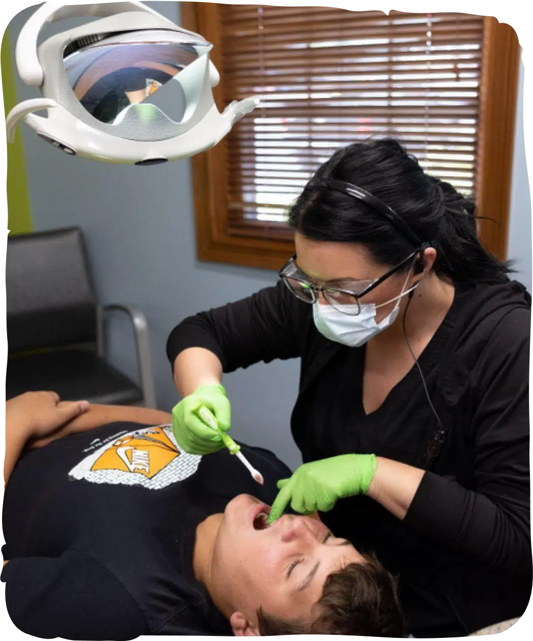 Pediatric dentist in green gloves examining young patient's teeth under overhead light in dental office.